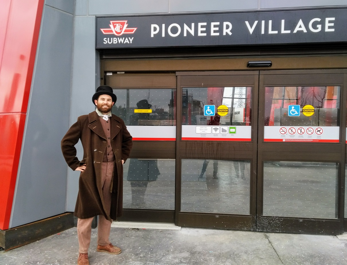 A man in 19th century clothes stands in front of the TTC Pioneer Village subway station